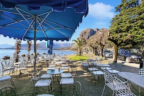 Un café al aire libre con mesas y sillas, un paraguas azul, y vistas al mar y a las montañas.