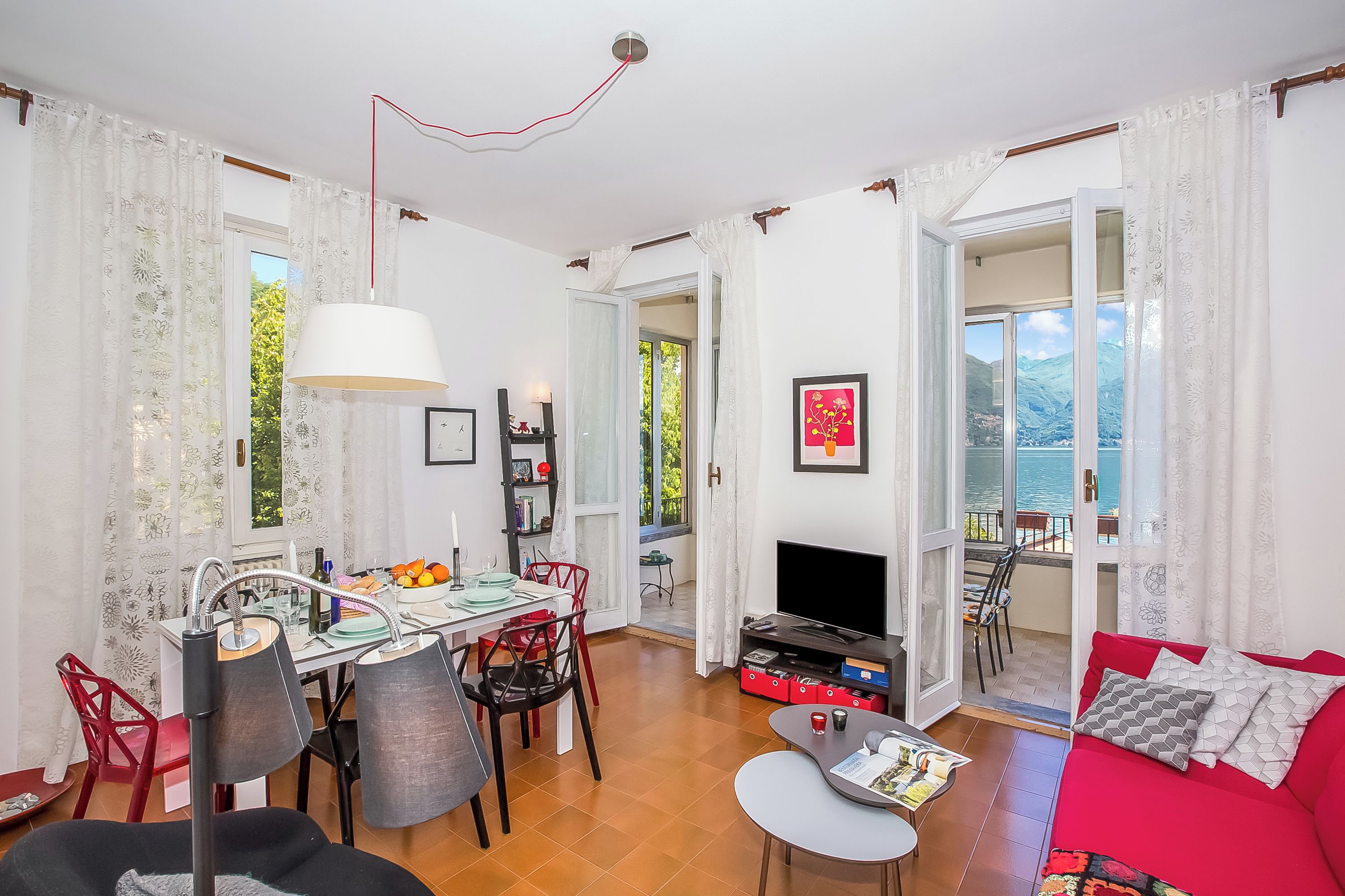 A dining area with a table, chairs, and a TV. There is a red sofa and a window with a view.
