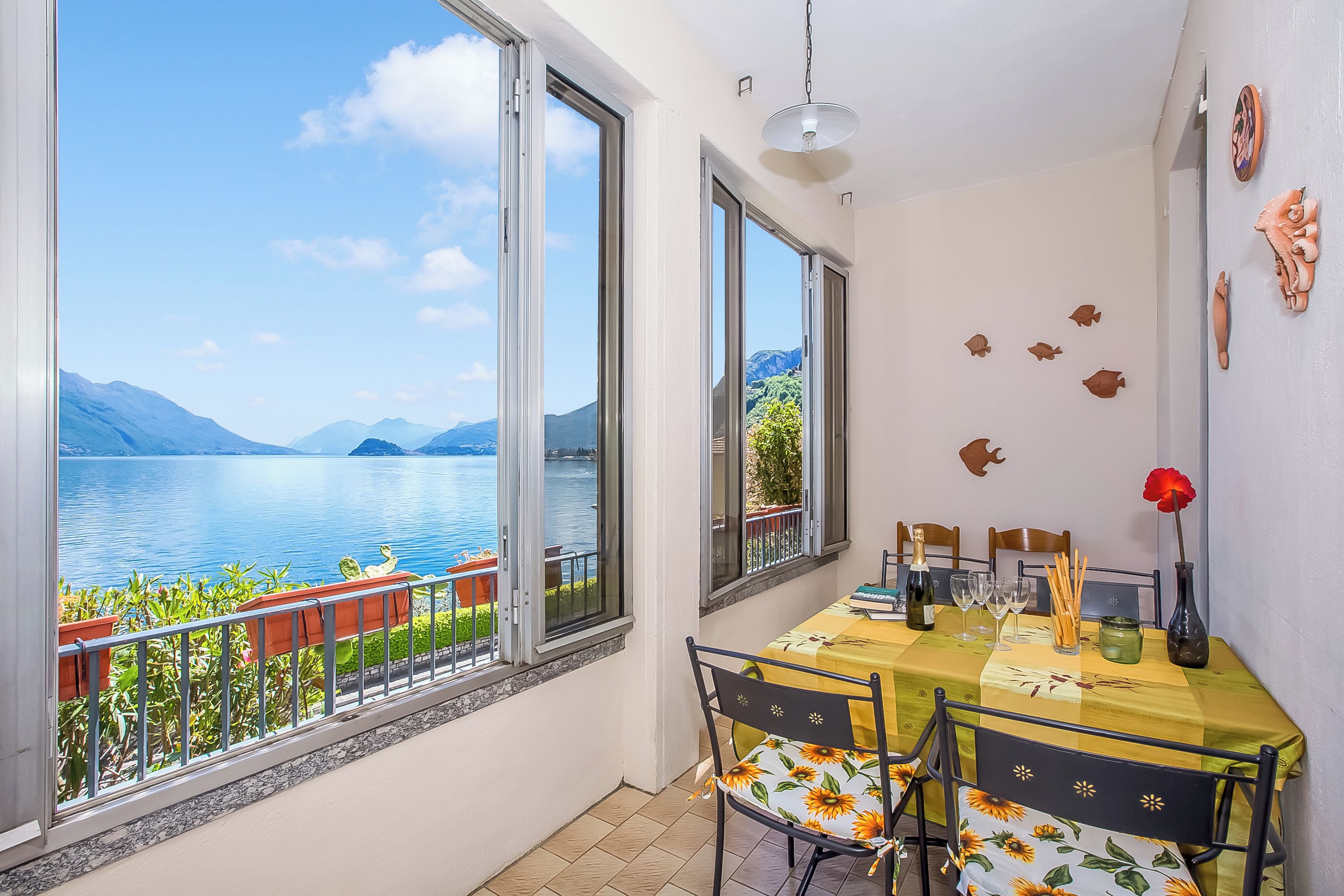A balcony with a view of a lake and mountains, a dining table with chairs, and a vase with a red flower.
