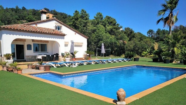 Une piscine entourée de chaises longues, un bâtiment blanc avec un toit en tuiles et, en arrière-plan, une végétation luxuriante.