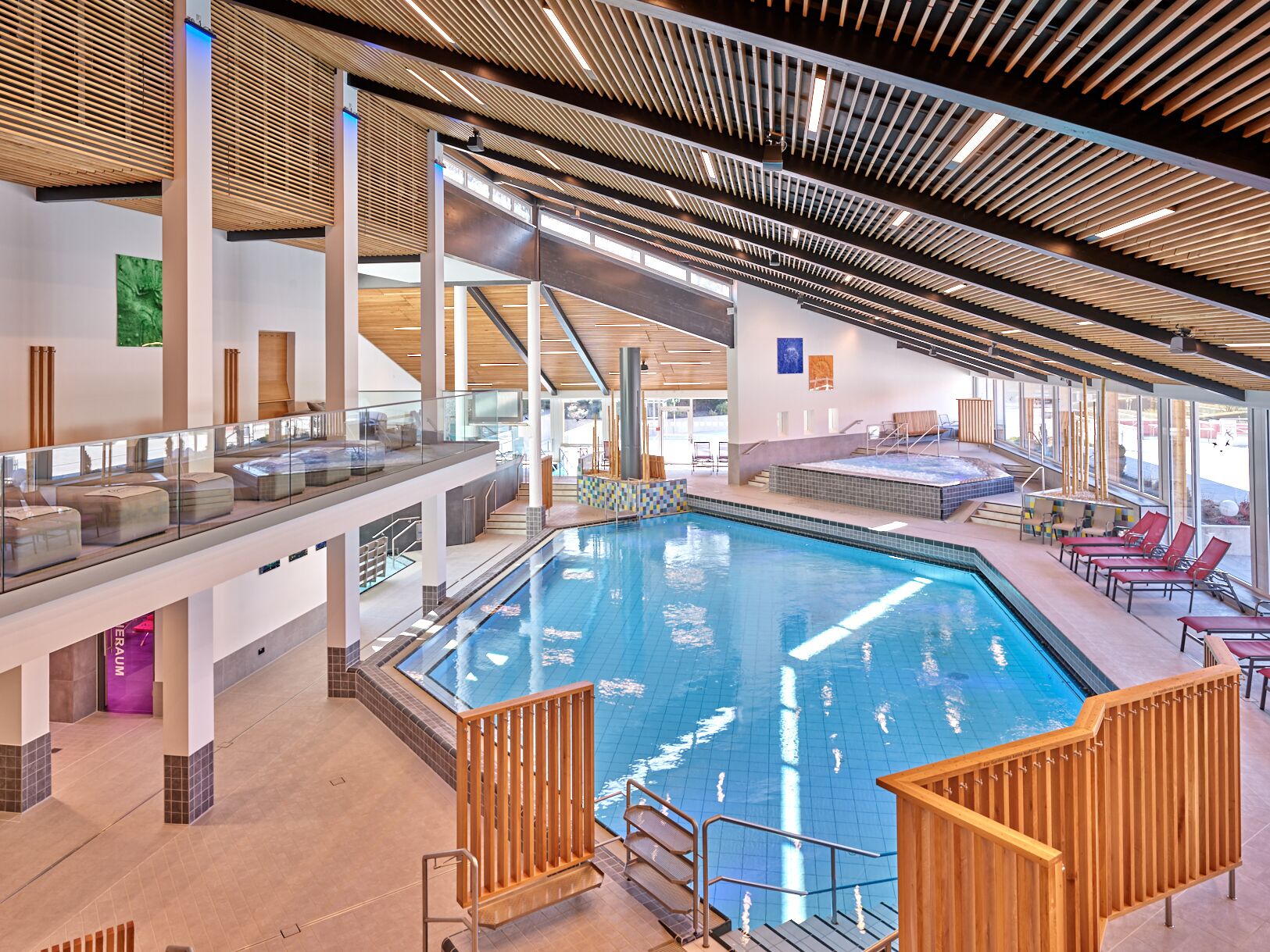 An indoor swimming pool with a glass balustrade, wooden deck, and lounge chairs.