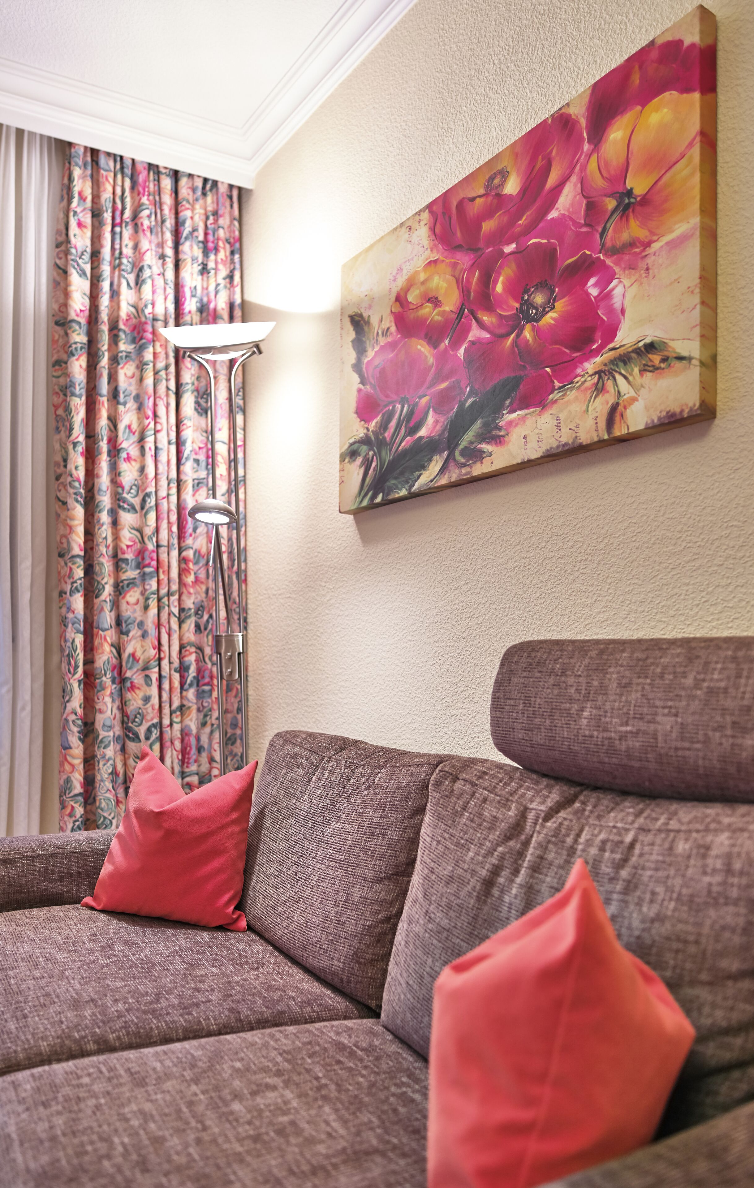 A living room with a brown sofa, red cushions, a floor lamp, and a floral painting on the wall.