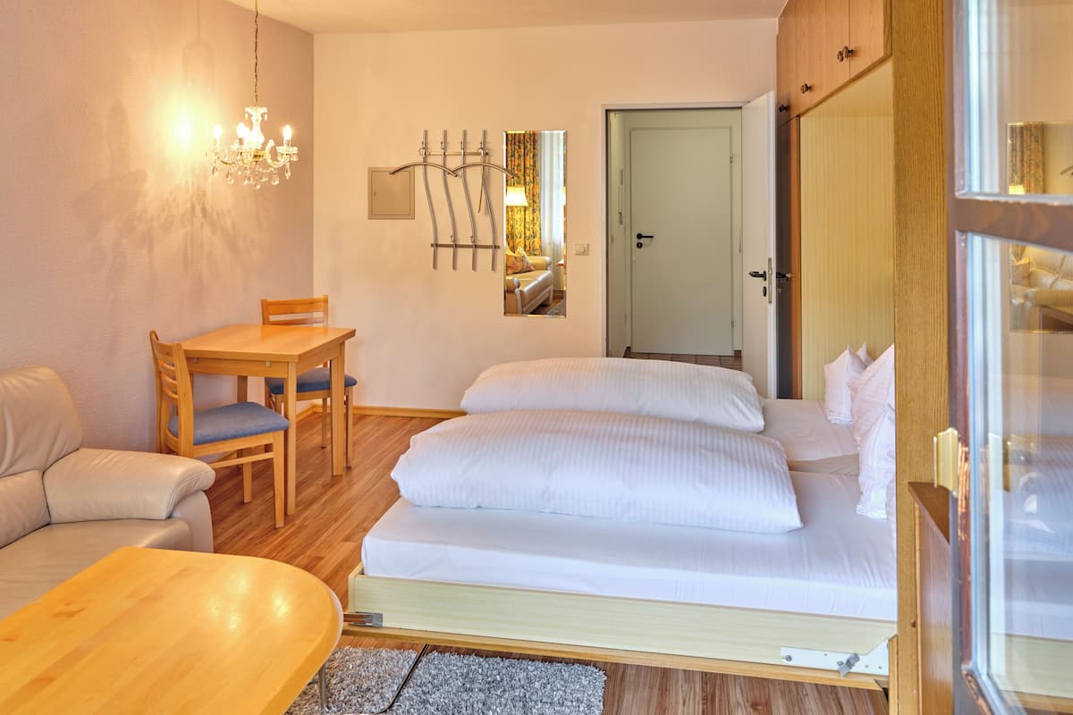 A compact hotel room with a bed, a wooden table, a chair, a sofa, and a chandelier.