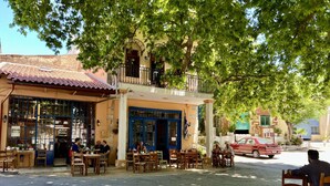 An outdoor café with tables and chairs under a tree, a building with a blue door, and a red car parked on the street. - Villa Galania (Armeni)