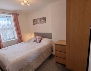 A bedroom with a bed, a nightstand, a wooden wardrobe, a window with curtains, and a wall-mounted artwork.