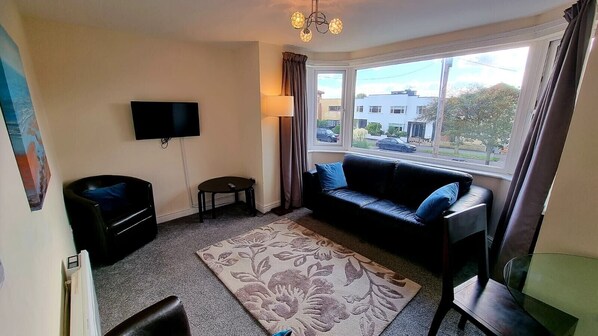 A living room with a sofa, a TV, a lamp, a small table, and a window with a view of buildings and trees.