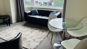 Ett modernt vardagsrum med en svart lädersoffa, ett glasbord och vita stolar. En mönstrad matta ligger på golvet, och ett fönster ger utsikt över byggnaderna utanför.
