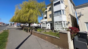 En flervåningsbyggnad med bostäder, med ett stort träd framför, en gata med bilar och en klarblå himmel.
