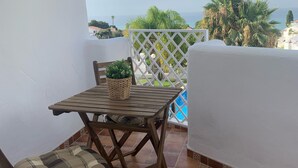 Uma varanda com mesa e cadeira de madeira, um cesto trançado e vista para o mar e palmeiras.