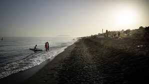 Uma praia com pessoas caminhando e paradas perto da água, céu claro e montanhas ao longe.