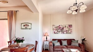 Un salón con una mesa redonda de comedor, dos sillas de madera, un sofá con cojines estampados, una mesita de centro, una lámpara y un cuadro floral en la pared.