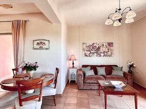 A living room with a round dining table, two wooden chairs, a sofa with patterned cushions, a coffee table, a lamp, and a floral painting on the wall.