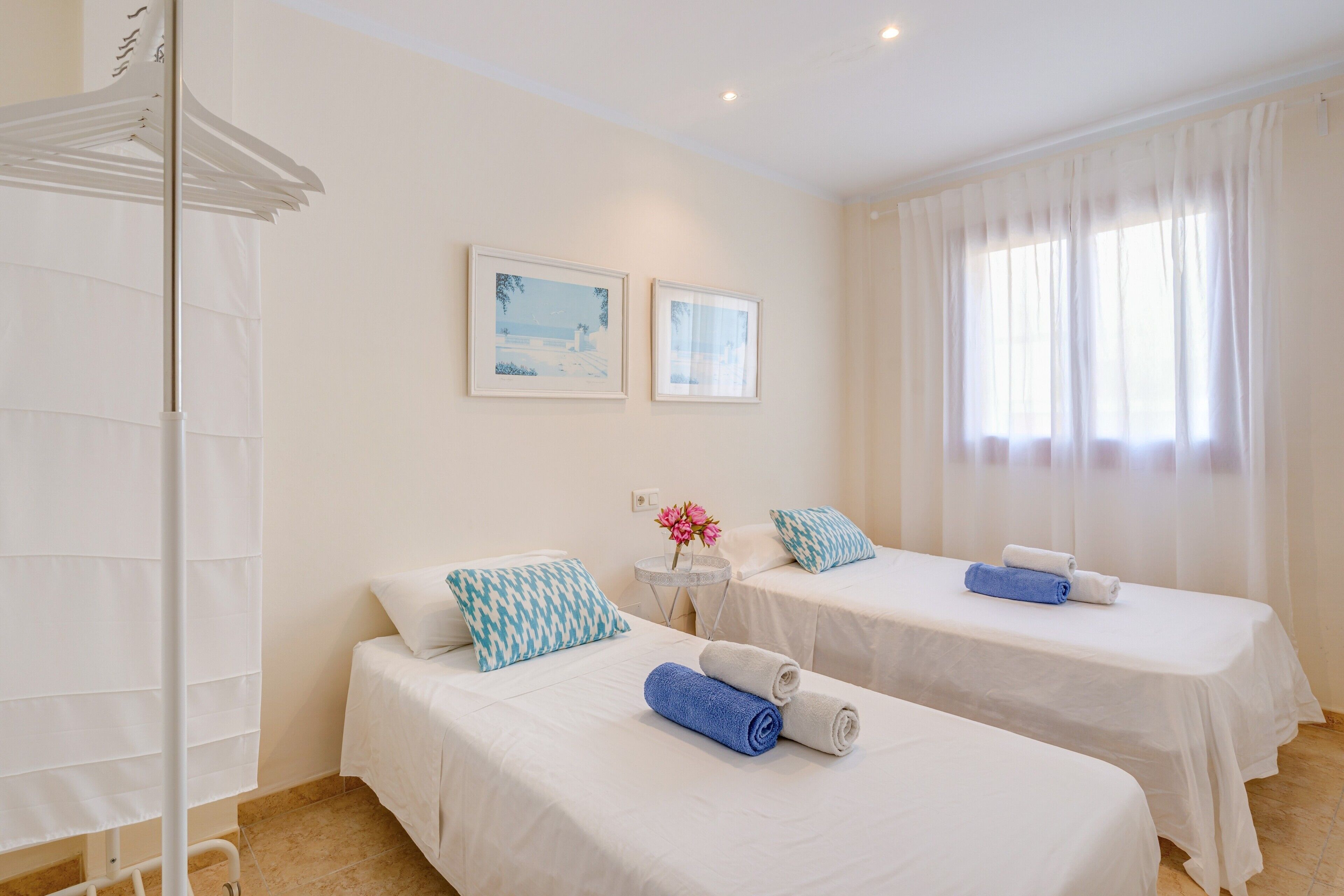 Dos camas individuales con ropa de cama blanca y toallas azules, una mesita con un florero y un armario blanco.