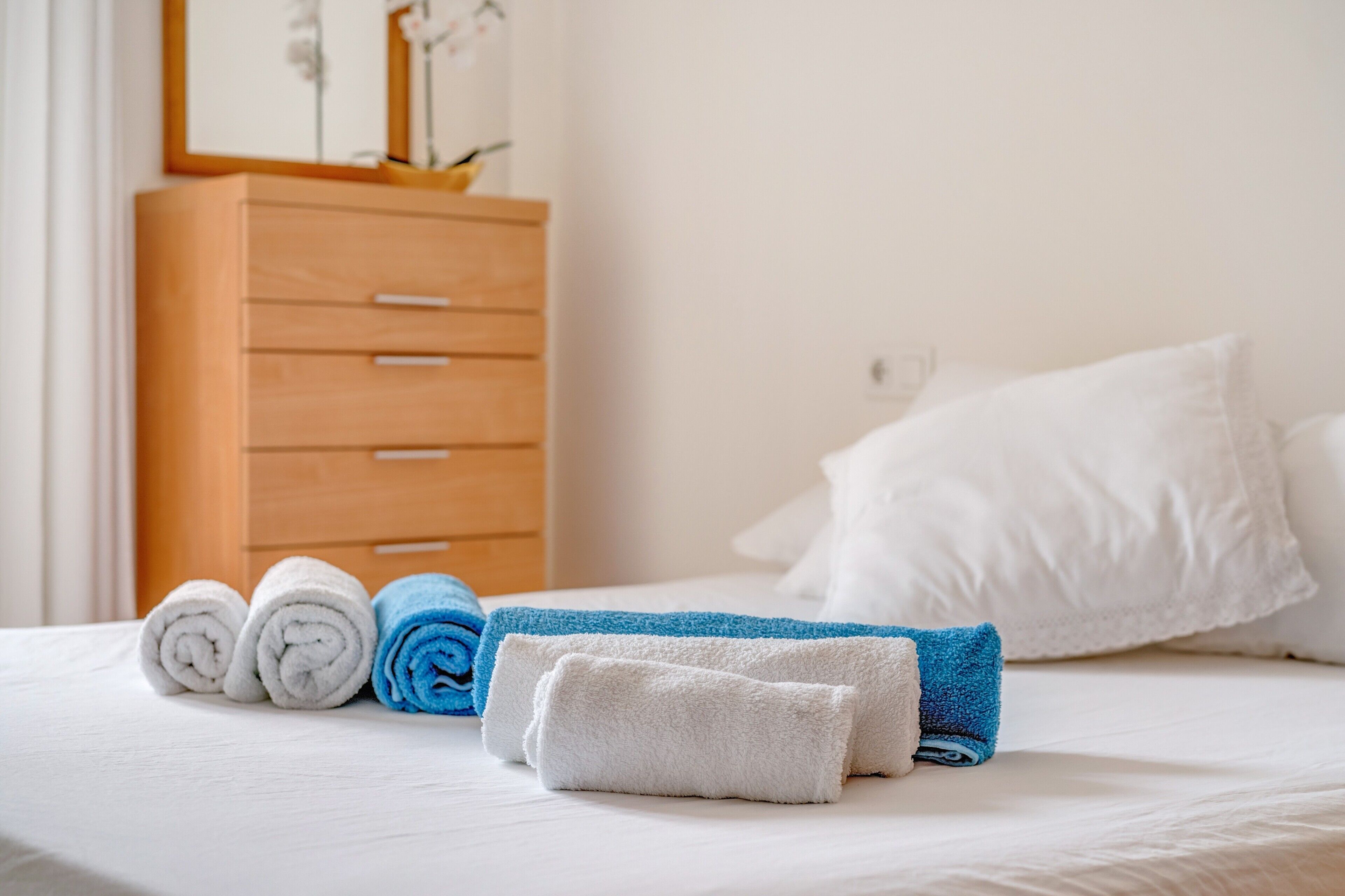 A neatly made bed with rolled towels and a wooden dresser in a hotel room.