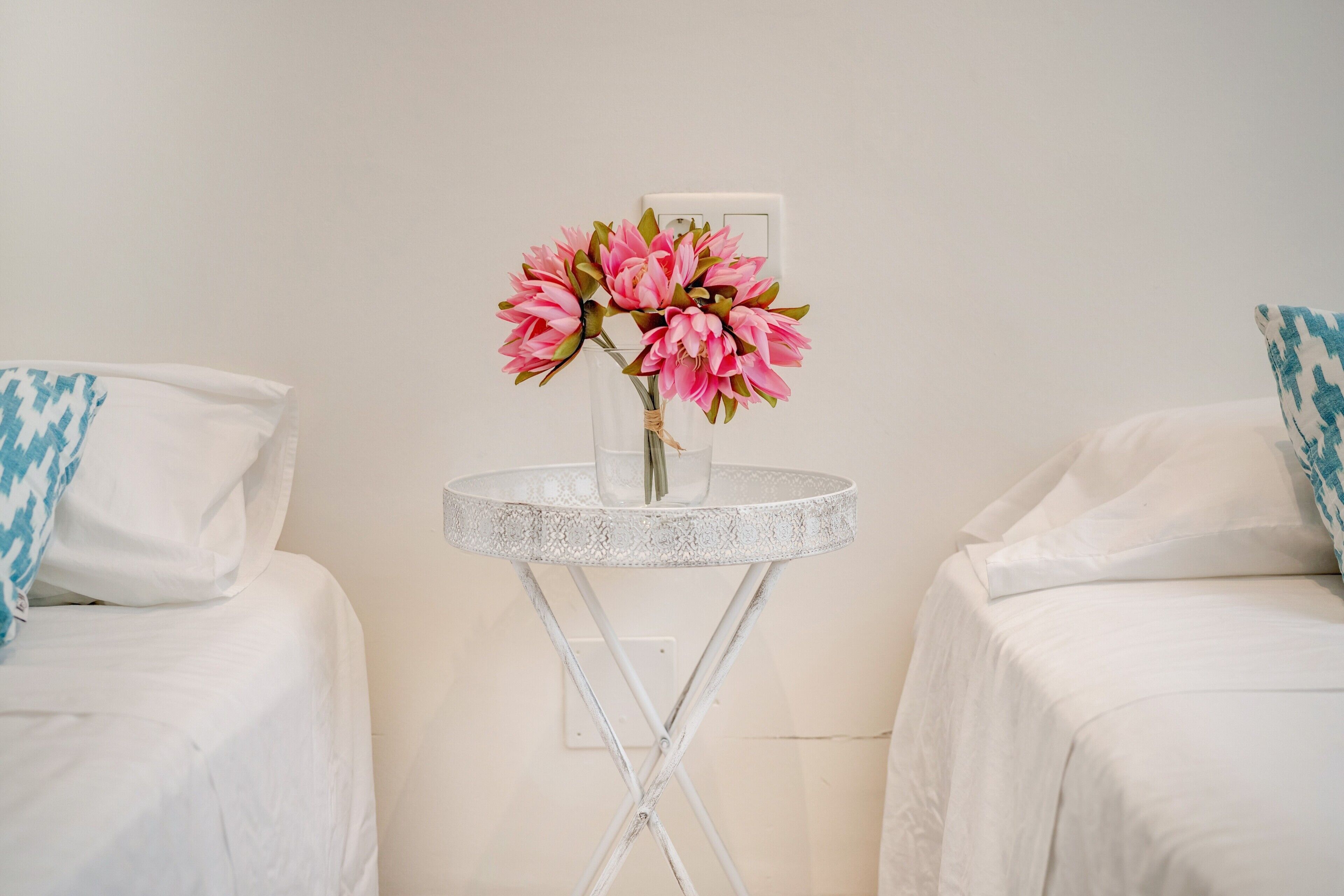 Une chambre avec deux lits, une petite table ronde ornée d’un vase de fleurs et un mur blanc.