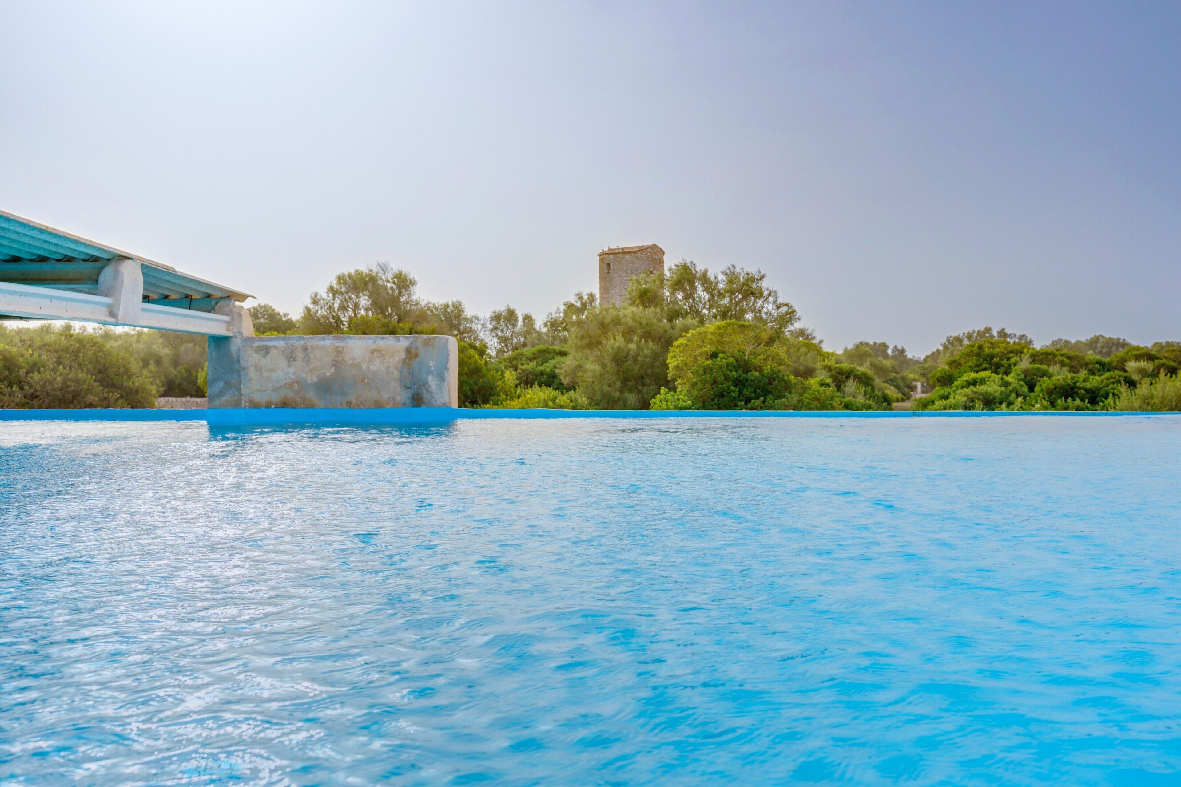 A swimming pool with a clear blue water surface, a stone tower in the background, and a bridge with a blue railing.