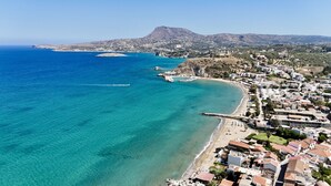 Vista aérea de un pueblo costero con aguas azules cristalinas, una playa de arena y un telón de fondo montañoso.
