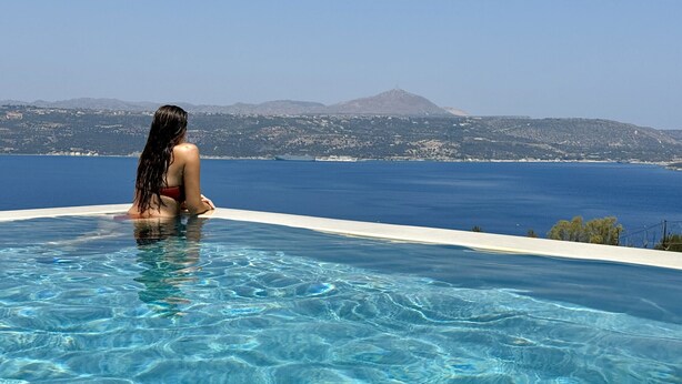 Una mujer sentada junto a una piscina cristalina con vistas a una montaña y al mar en la distancia.