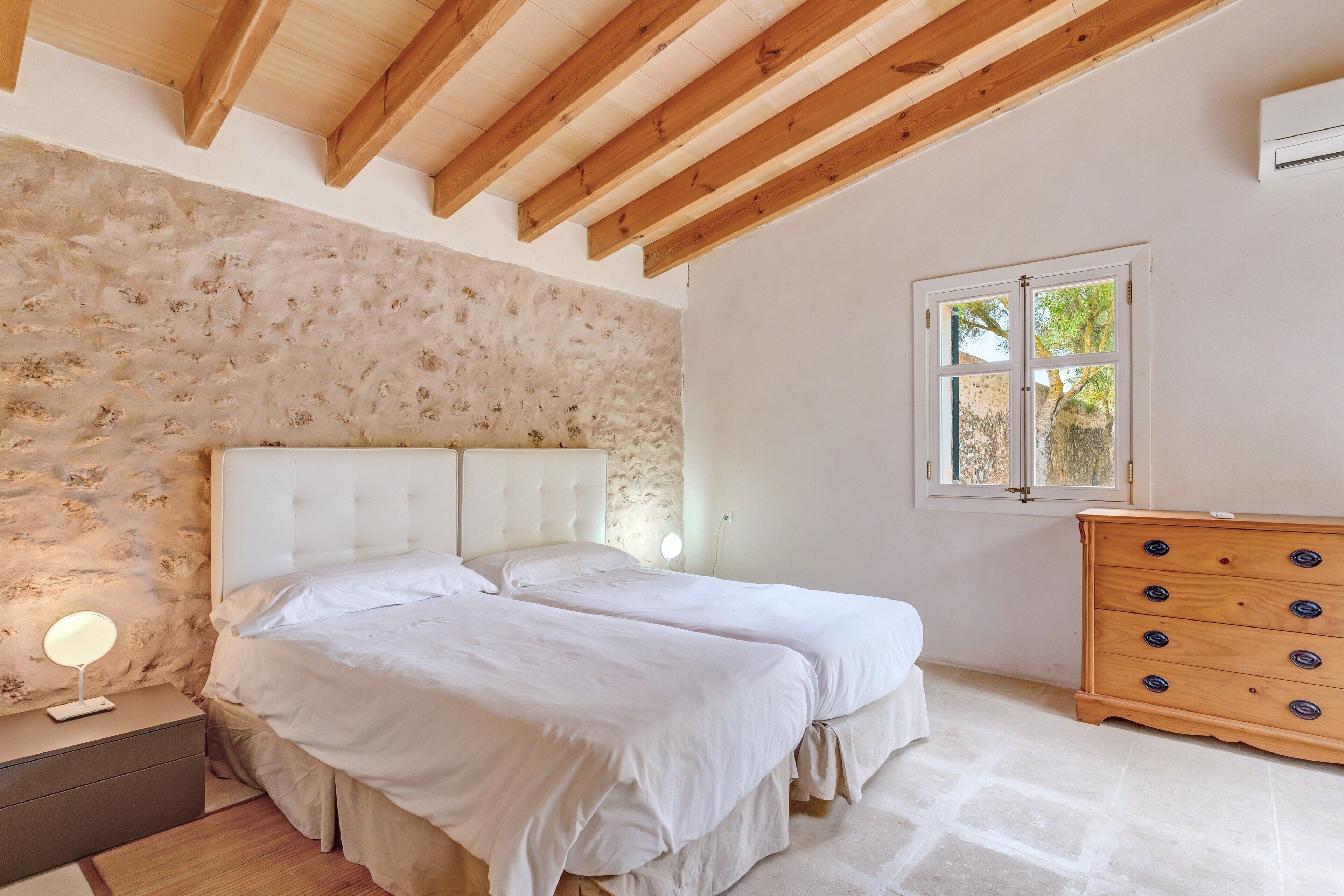 Ein Schlafzimmer mit Steinwand, Holzbalkendecke, einem Bett mit weißer Bettwäsche, einer Kommode mit mehreren Schubladen und einem Fenster mit Aussicht.