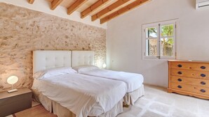 A bedroom with a stone wall, wooden ceiling beams, a bed with white bedding, a dresser with multiple drawers, and a window with a view.