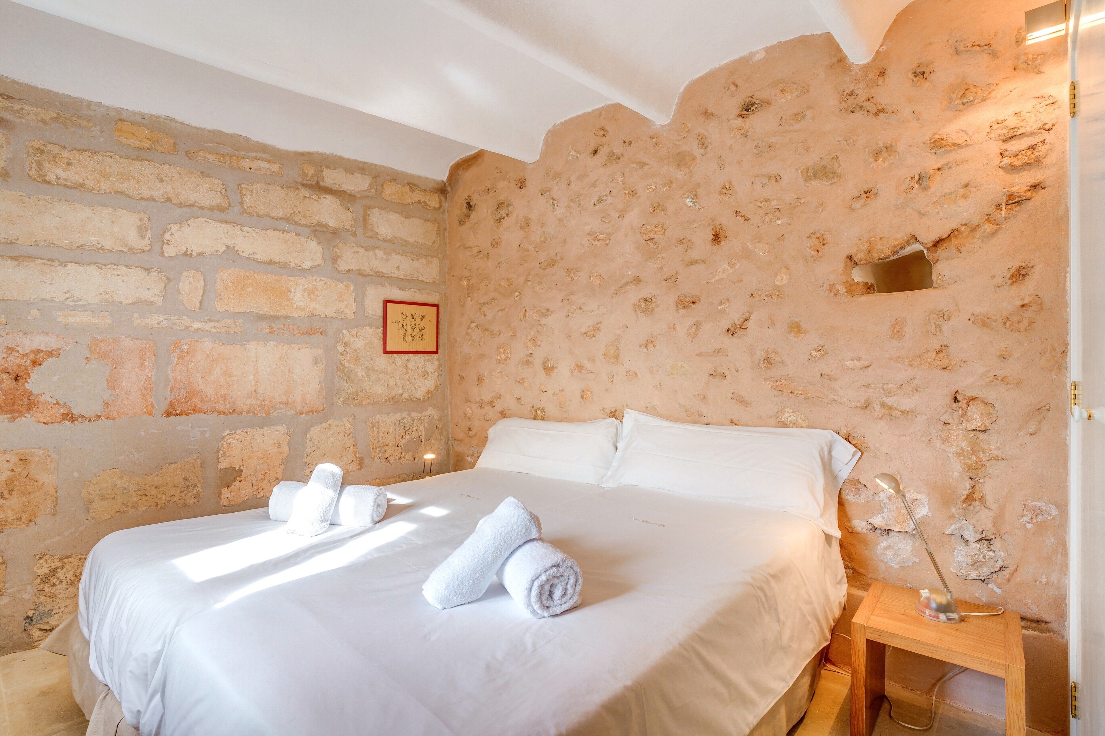 Une chambre avec un mur en pierre, un lit recouvert de draps blancs et une table de chevet en bois.