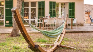 A hammock in a garden area with a wooden bench, a table with chairs, and a building with green shutters in the background.
