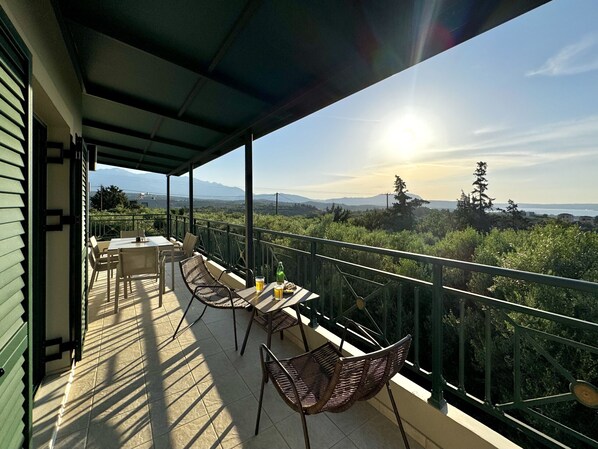 A balcony with a table and chairs, overlooking a landscape with trees and distant mountains. - Villa Nynemia (Almyrida)