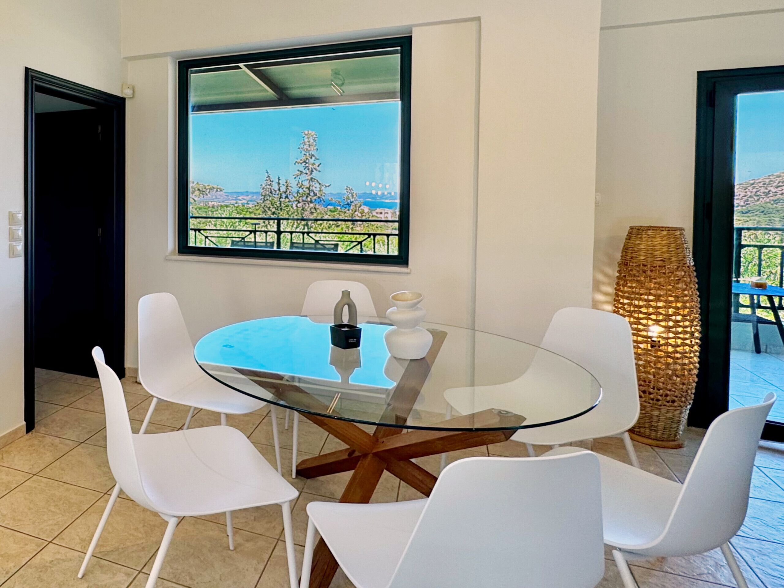 Een moderne eethoek met een glazen tafel, omringd door witte stoelen, een groot raam met uitzicht op groen en een rieten lamp.