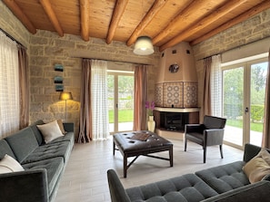 A cozy living room with a stone wall, wooden ceiling beams, a fireplace, and a large sliding glass door leading to a garden.