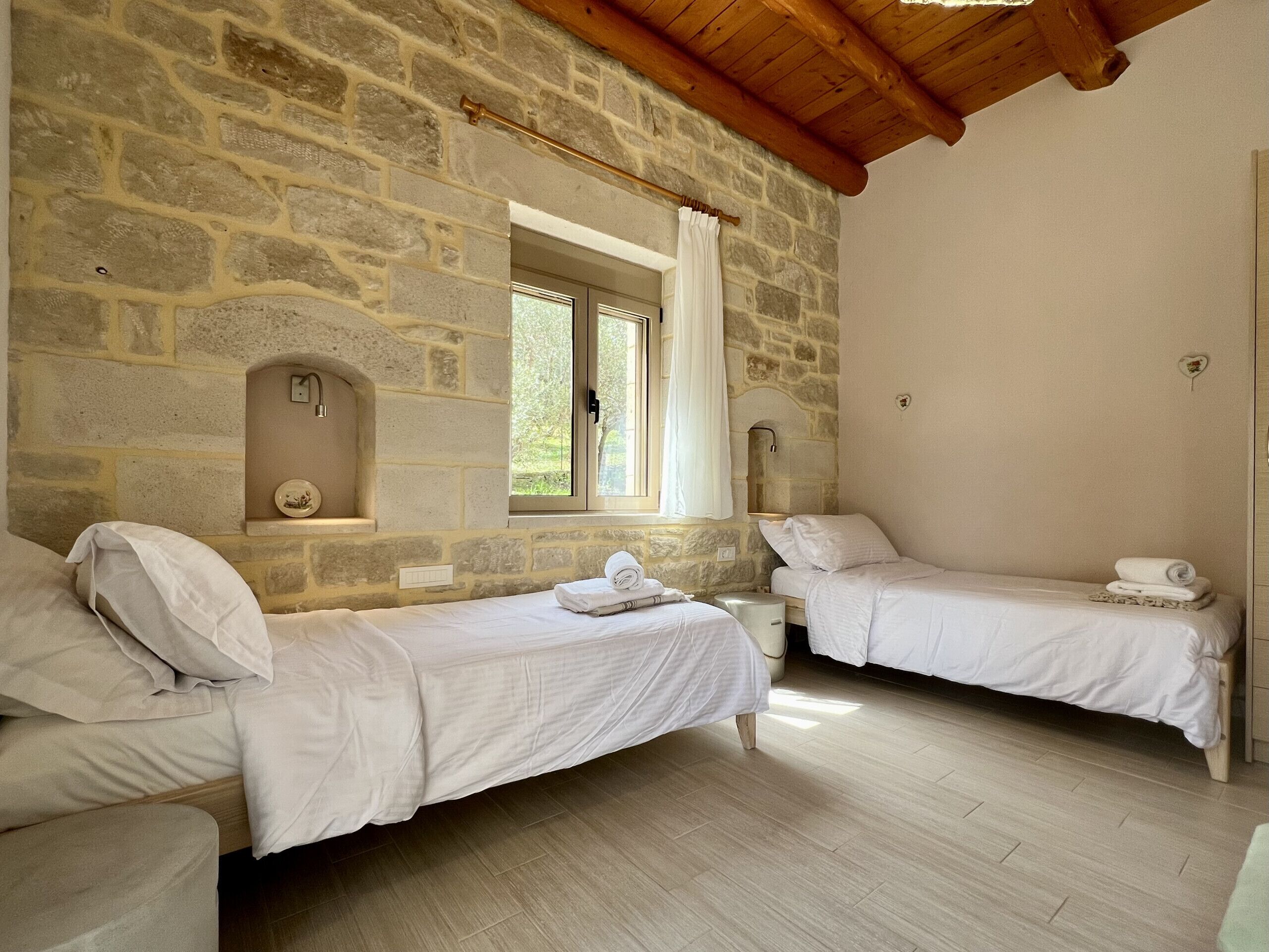 Une chambre avec deux lits simples, un mur en pierre, un plafond en bois, une fenêtre avec des rideaux et une petite étagère sur laquelle est posé un objet décoratif.