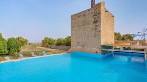 Una piscina con una estructura de piedra, sillones de descanso y una noria al fondo.