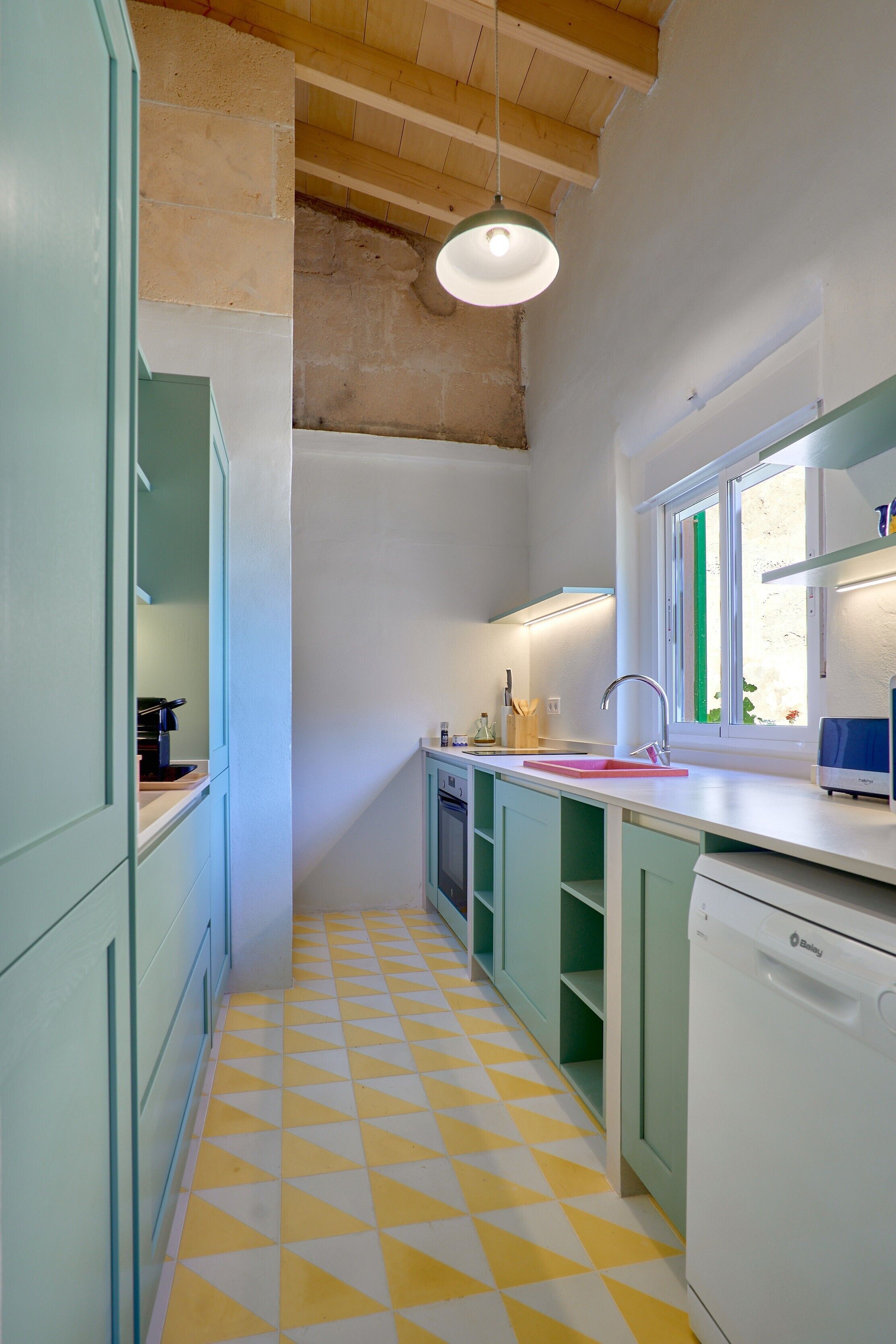 A kitchen with a yellow and white checkered floor, green cabinets, a white dishwasher, and a window with a view.