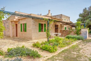 Un bâtiment en pierre avec des volets verts, un jardin où poussent différentes plantes et un mur en pierre.