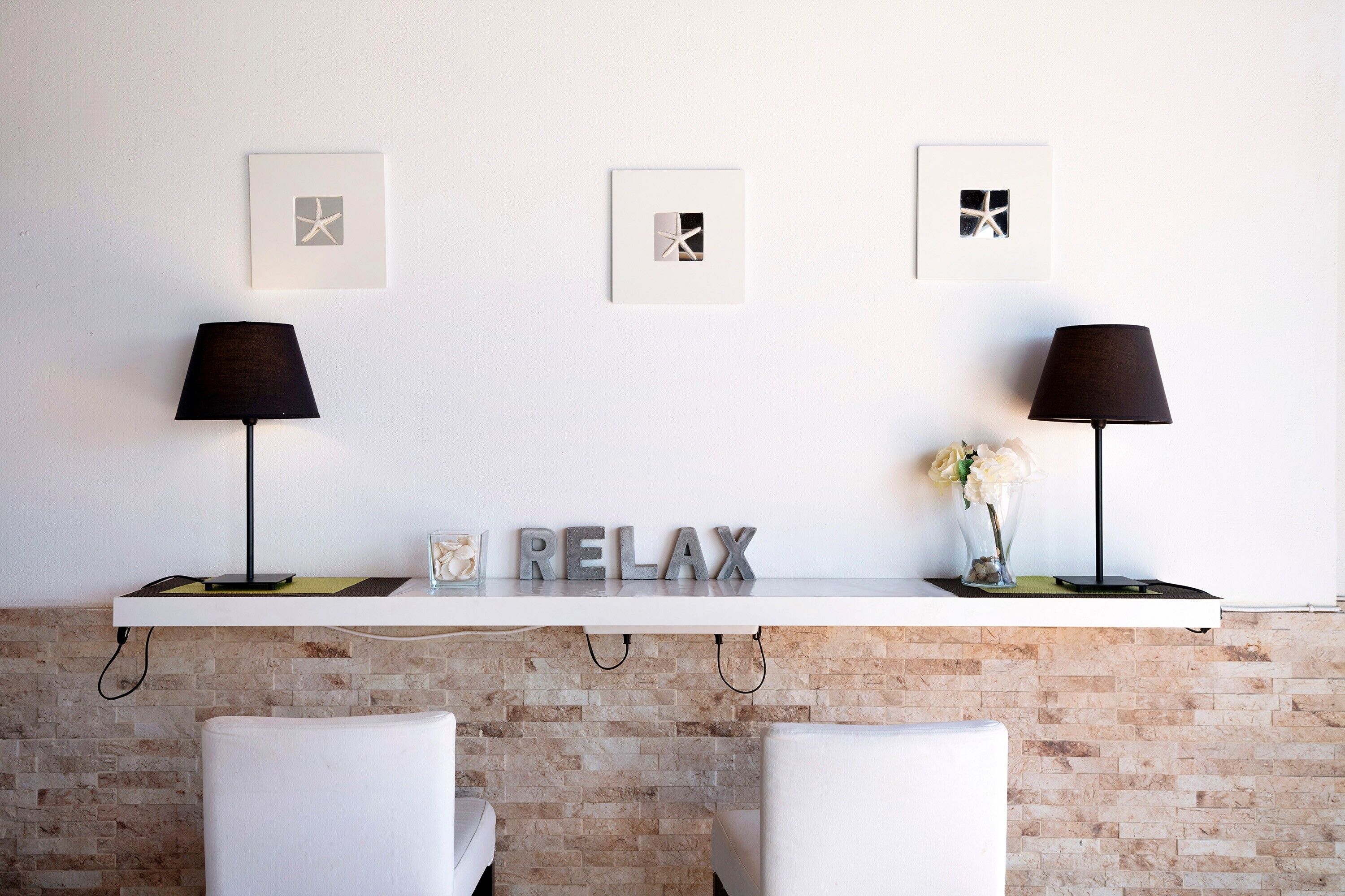 A modern room with a white brick wall, two white chairs, a white shelf with a 'RELAX' sign, and three framed pictures.