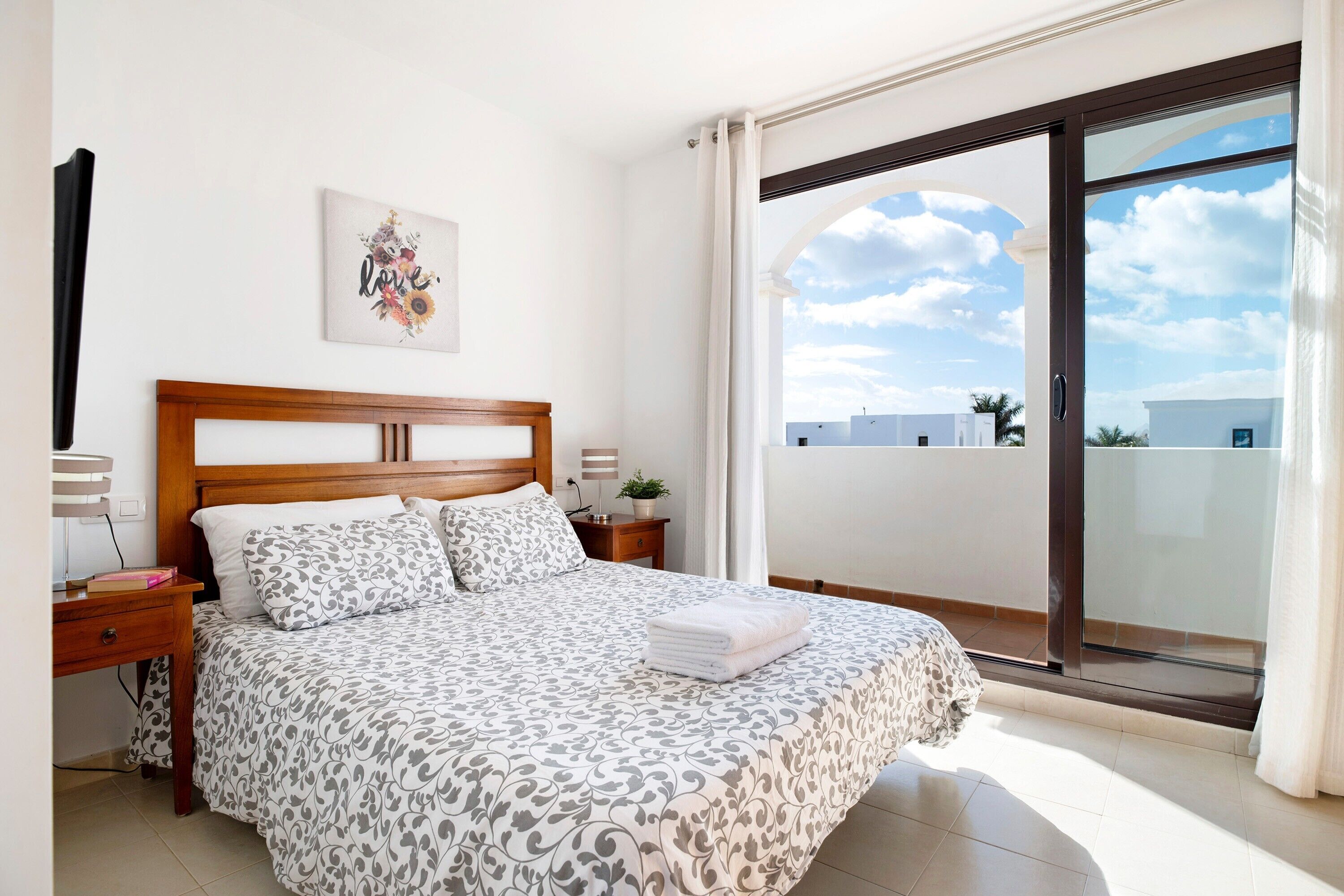 Un dormitorio con cama, mesita de noche, una obra de arte en la pared y un amplio ventanal corredero que da a un balcón.