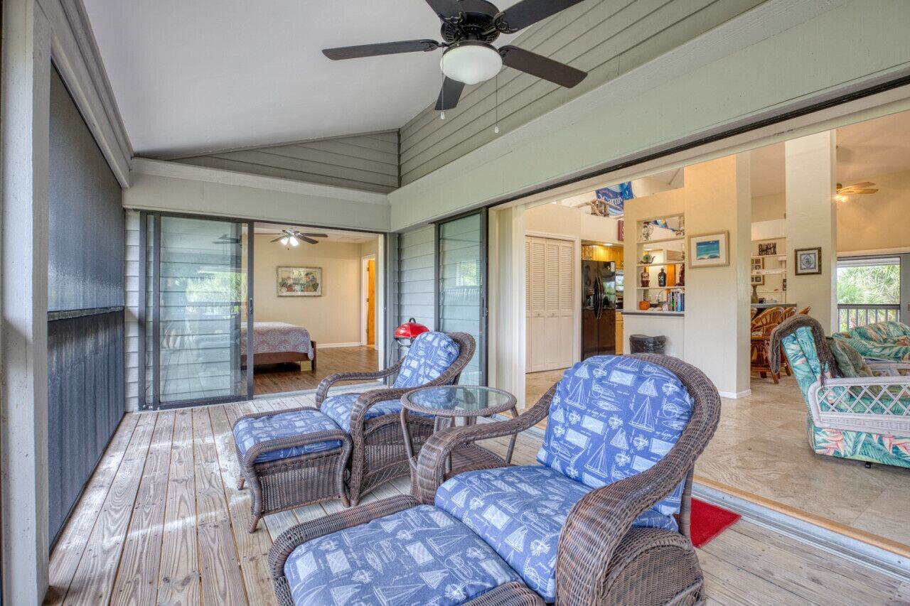 Une terrasse couverte, dotée de meubles en osier, d’un ventilateur de plafond et offrant une vue sur l’intérieur de la maison.