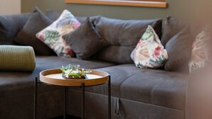 A sectional sofa with floral cushions, a small wooden side table with a glass and flowers, and a green cushion. - Apartment “Pritscheshof Balkon” with Mountain View, Balcony & Terrace (Mals)