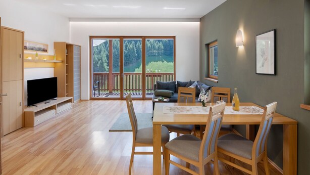 A spacious living area with a large window, wooden flooring, a dining table with chairs, a sofa, and a television. - Apartment “Pritscheshof Balkon” with Mountain View, Balcony & Terrace (Mals)