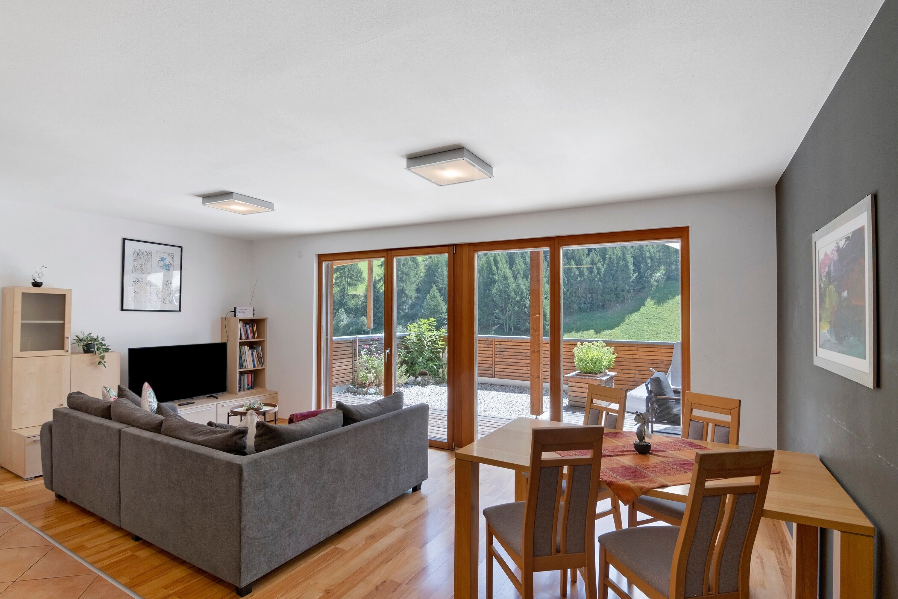 Ein modernes Wohnzimmer mit grauem Sofa, Fernseher und Bücherregal. Es gibt einen Essbereich mit Tisch und Stühlen. Große Fenster bieten Blick auf einen Garten und Bäume.
