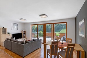 Un salon moderne avec un canapé gris, un téléviseur et une bibliothèque. On y trouve également un coin repas avec une table et des chaises. De grandes fenêtres offrent une vue sur un jardin et des arbres.