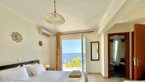 A hotel room with a bed, a nightstand, a lamp, a view of the sea, and a bathroom visible through an open door. - Villa Sakos (Skala)