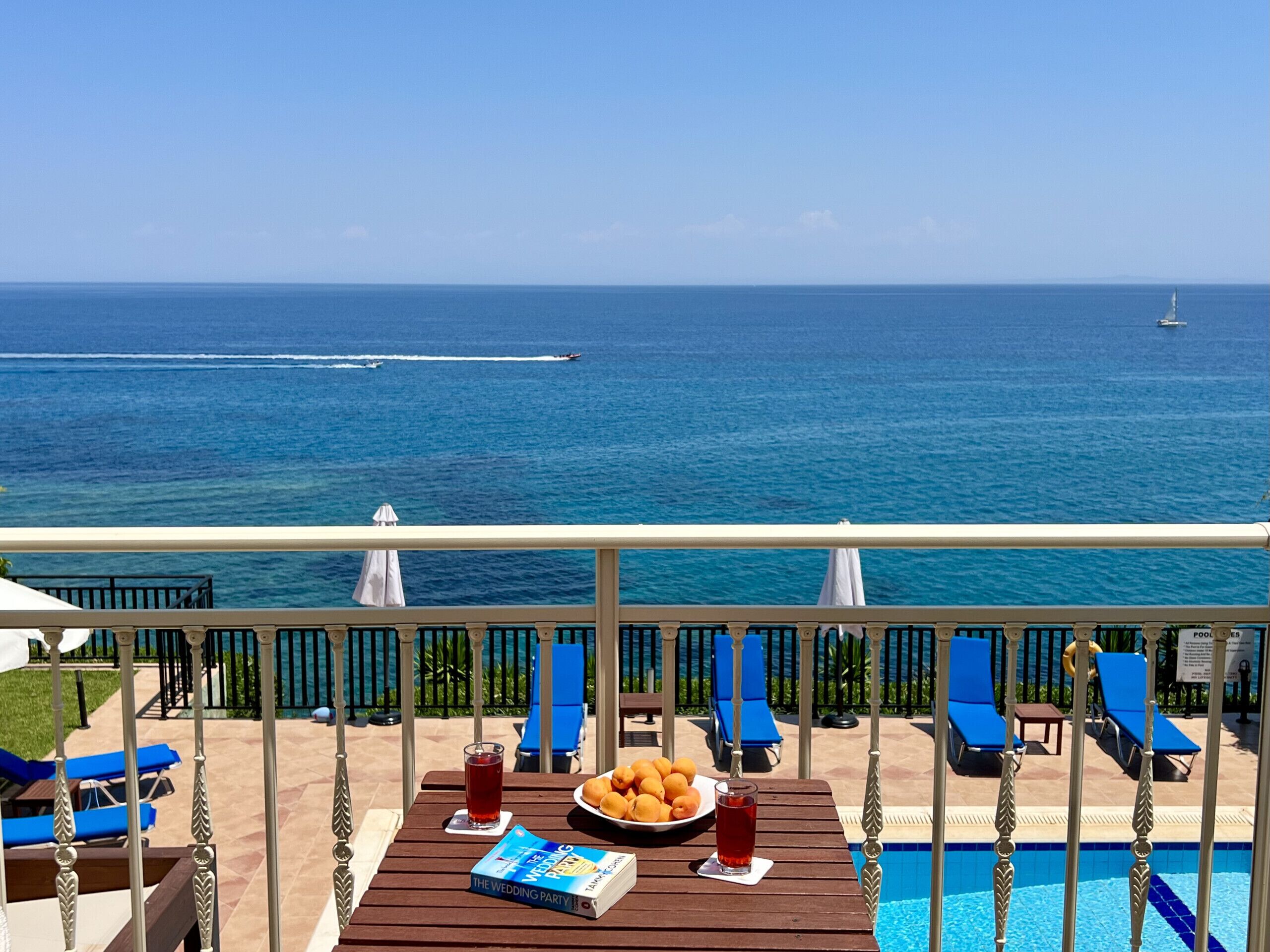 A poolside view with a view of the ocean, a table with a book and drinks, and lounge chairs.