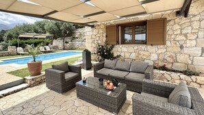 Un patio exterior con mobiliario de mimbre, una pared de piedra, una piscina y una zona cubierta con vistas a montañas.