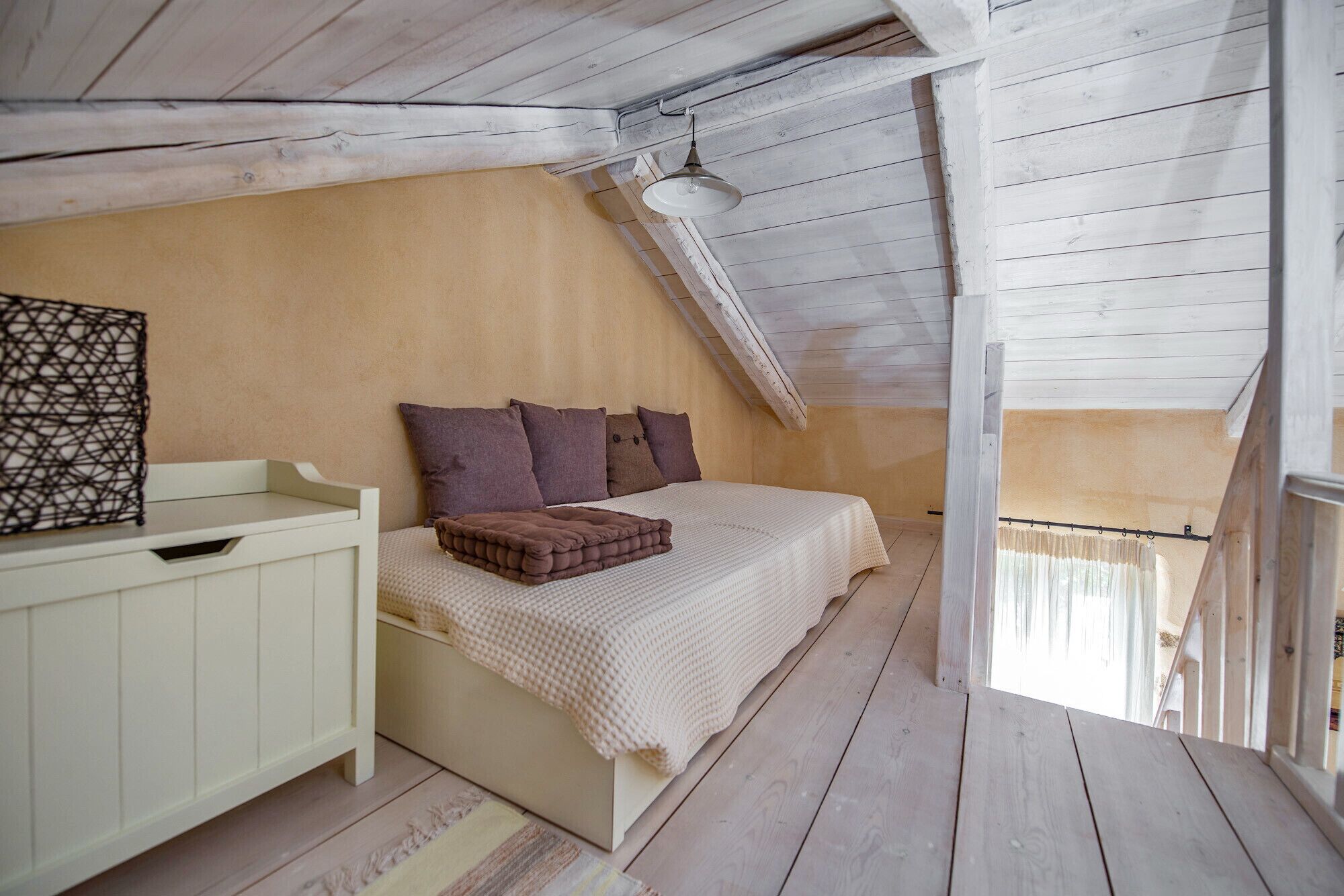 Ein Schlafzimmer mit geneigter Decke, einem Bett mit einer beigefarbenen und braunen Decke, einem weißen Nachttisch und einem Fenster mit Vorhang.