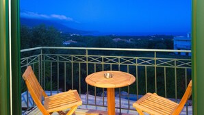 Un balcone con due sedie di legno e un tavolino rotondo, con vista panoramica su una zona costiera con edifici e vegetazione.