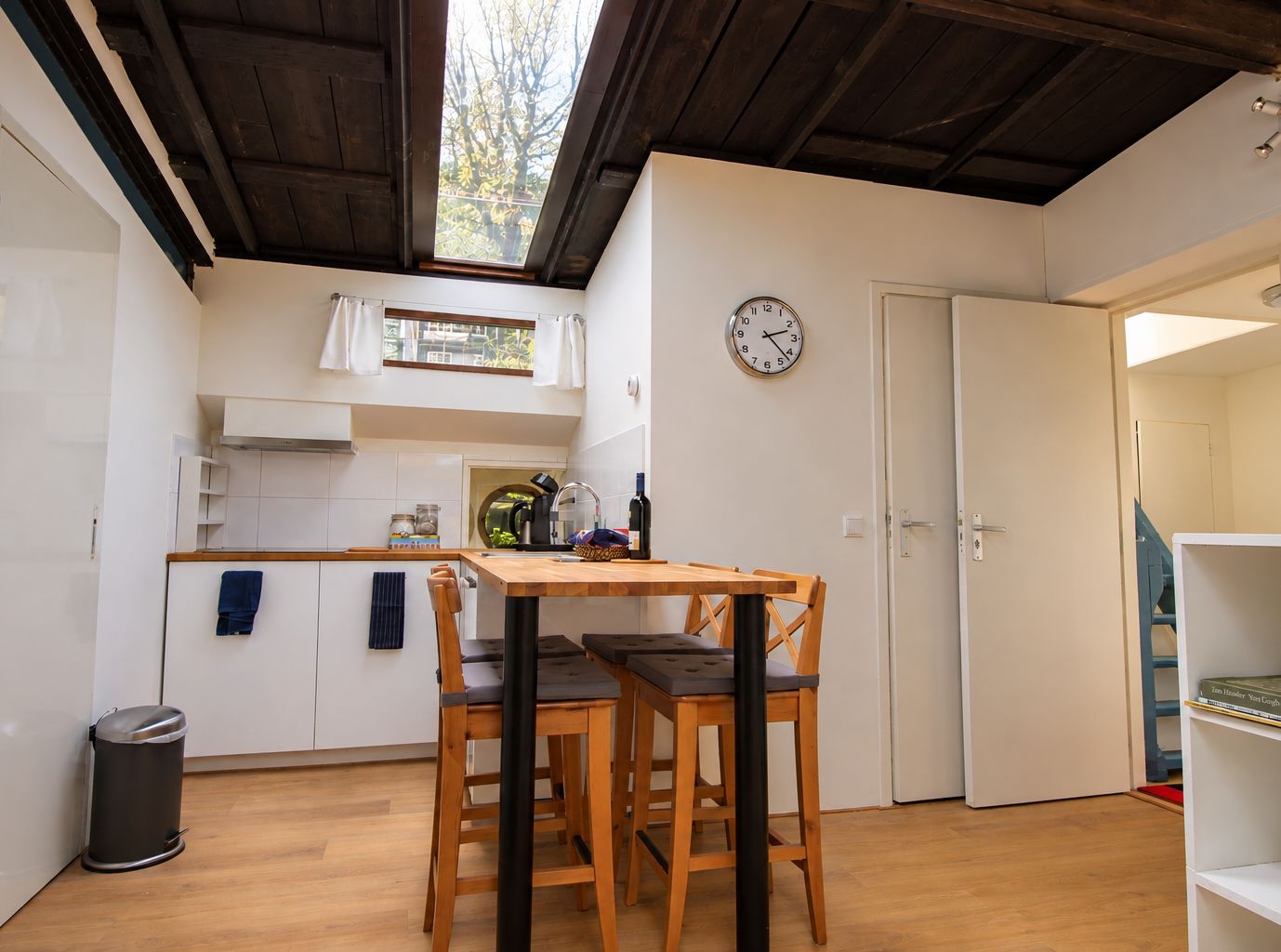A compact kitchen with a dining table, chairs, a clock, and a view of the outdoors through a window.