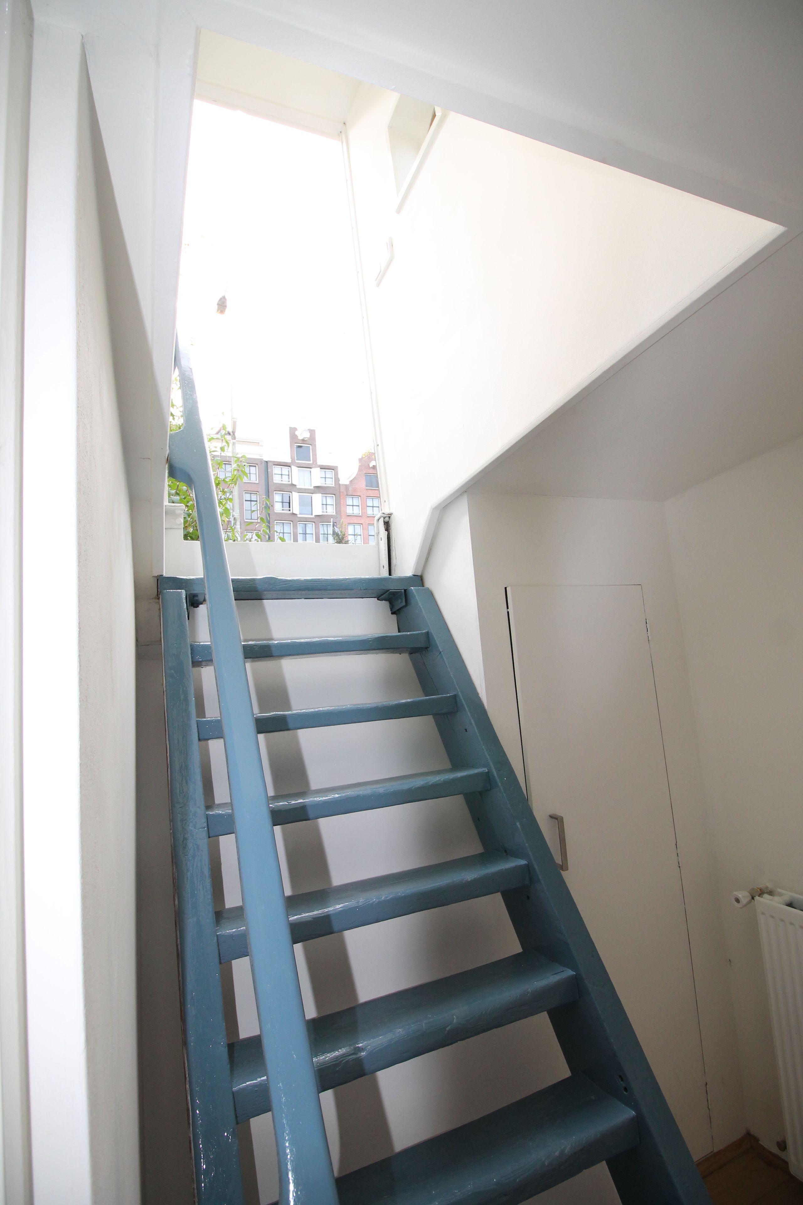 Eine blaue Holztreppe führt zu einer weißen Tür, mit Blick auf Gebäude im Hintergrund.