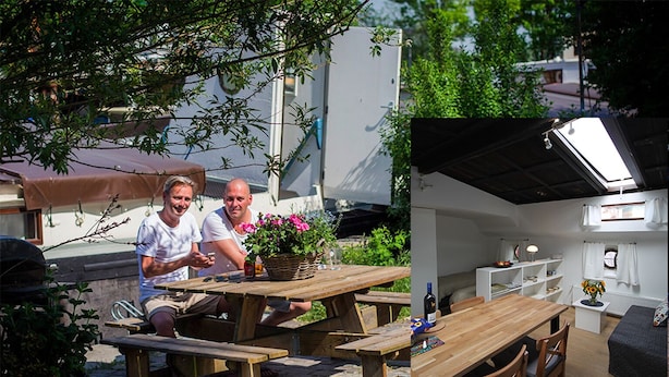 Un patio al aire libre con mesas y bancos de madera, un cesto de flores y vistas a una zona residencial con árboles y un edificio blanco al fondo.