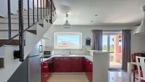 A modern kitchen with red cabinets, a white countertop, and a built-in oven. There is a window with a view of the outside, and a staircase leading to an upper floor. - Villa Pascal (Peruški)