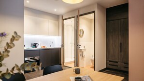 Een moderne hotelkamer met een houten eettafel, een koffieapparaat en zicht op de keuken en badkamer door een open deur.